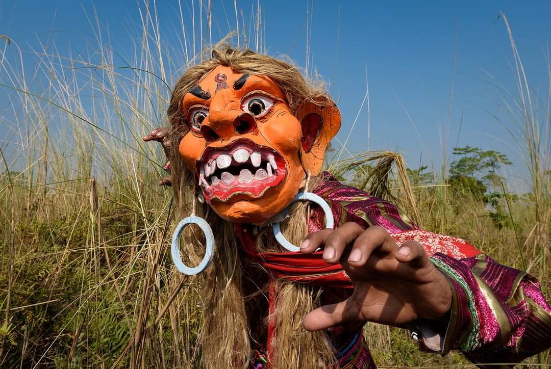 Bhaona performer wearing a ferocious orange demon mask in an open field
