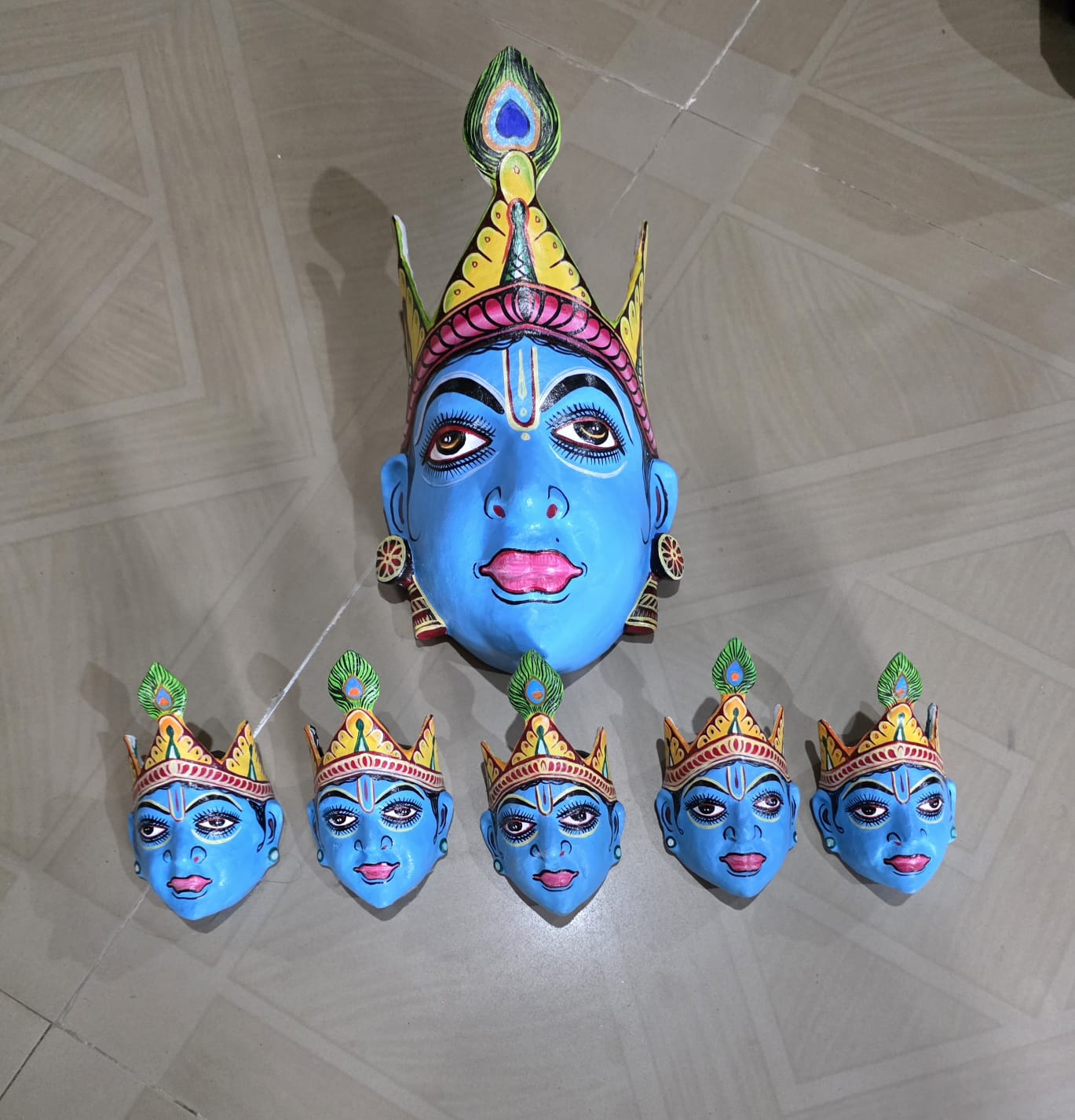 A large blue Krishna mask with a peacock-feather crown beside five smaller blue masks